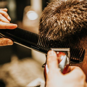 A cutting comb held flat against the back of a client's head while clippers run along it. Photographed from behind; no face is in frame.