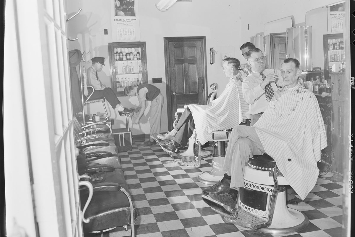 Barbers in white coats cutting customers' hair at a row of chairs in a 1940s works barbershop.