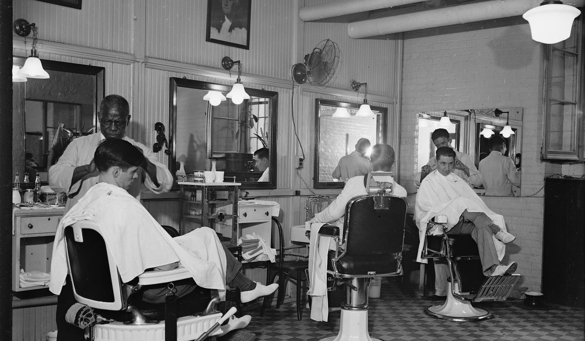A long row of barber chairs with barbers at work in a 1930s office-building barbershop.