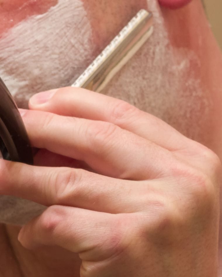 A straight razor drawn across a lathered cheek while the fingers of the other hand stretch the skin.