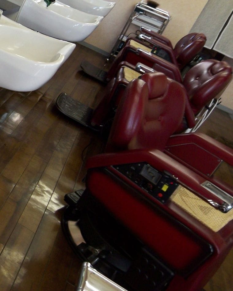 A row of red leather barber chairs facing white basins along a narrow shop with a wooden floor.