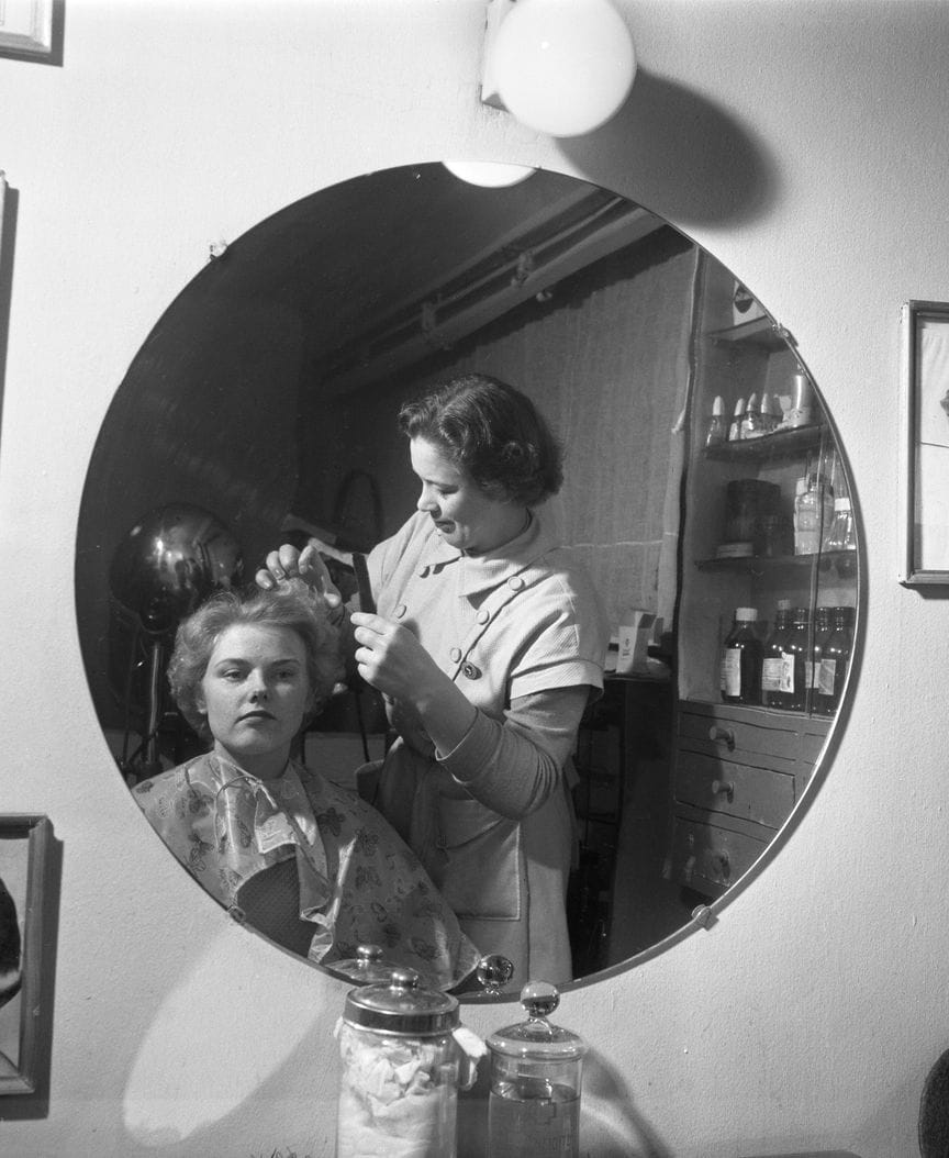 A black-and-white photograph of a hairdresser setting a client's hair, both reflected in a round salon mirror.