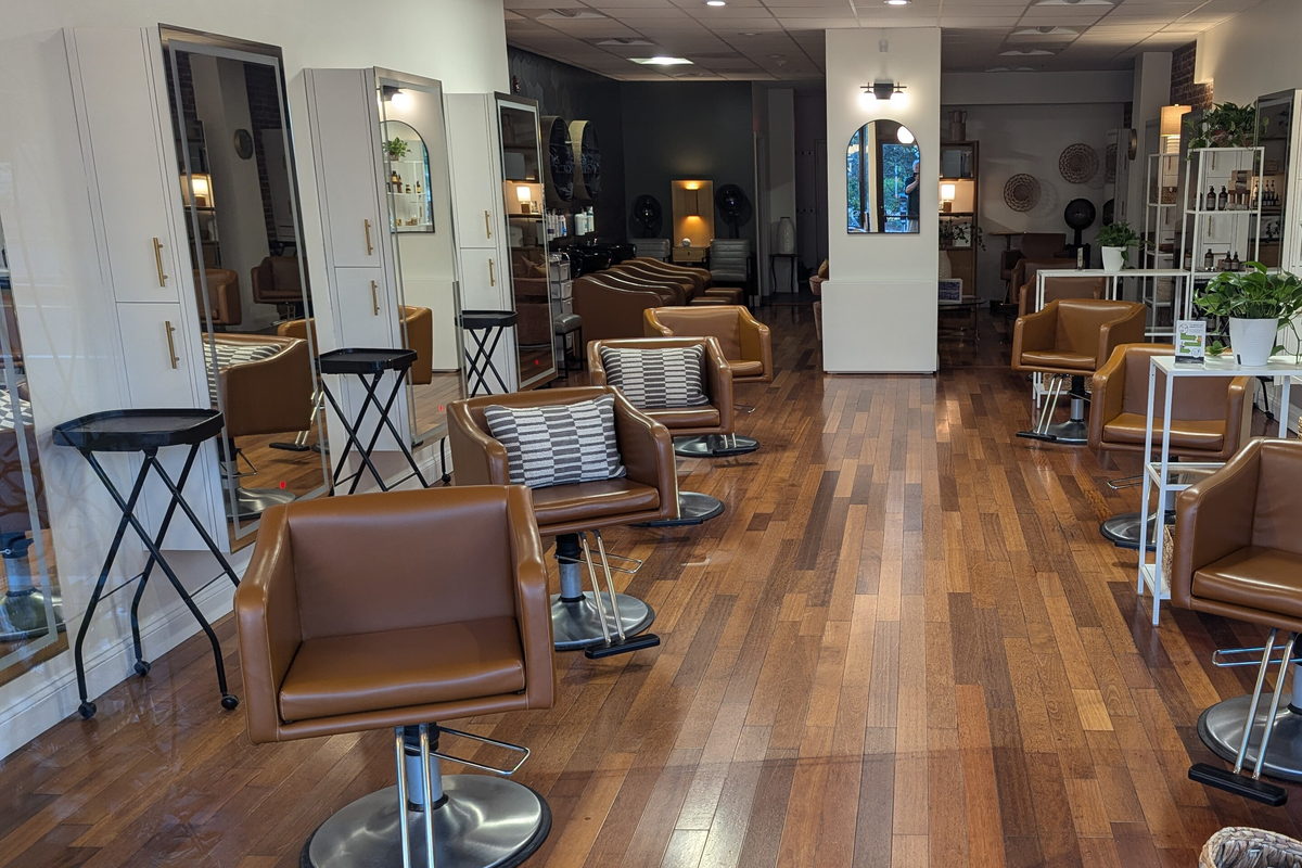 A long salon room lined with mirrors, tan styling chairs and plants above a pale wood floor.