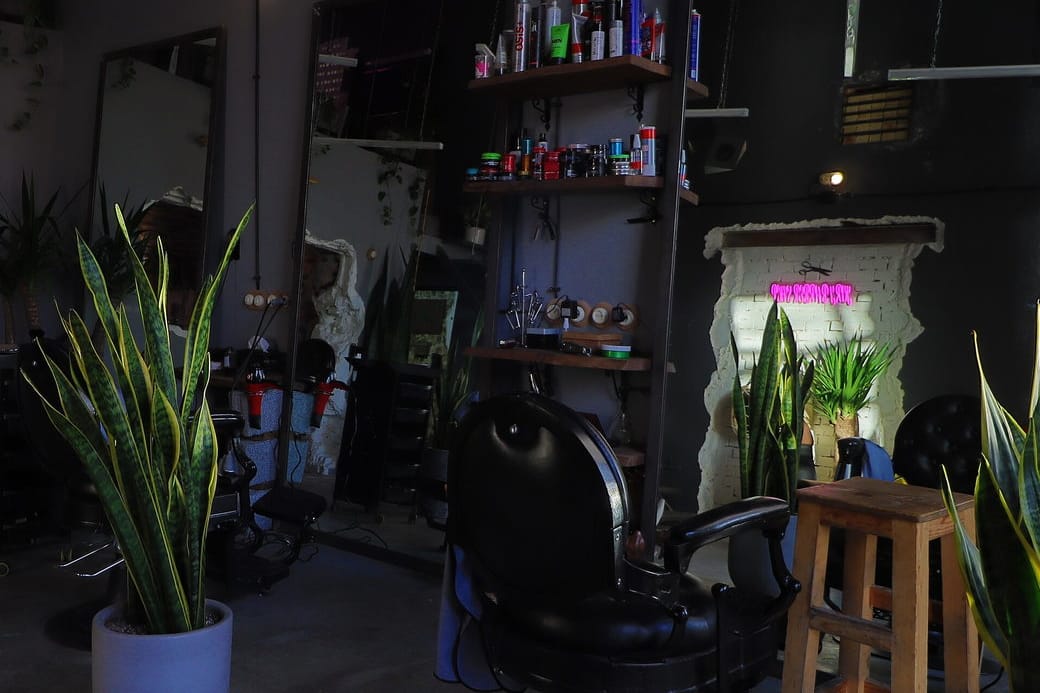 An empty styling chair beside a wooden stool and plants in a dim, uncluttered salon room.