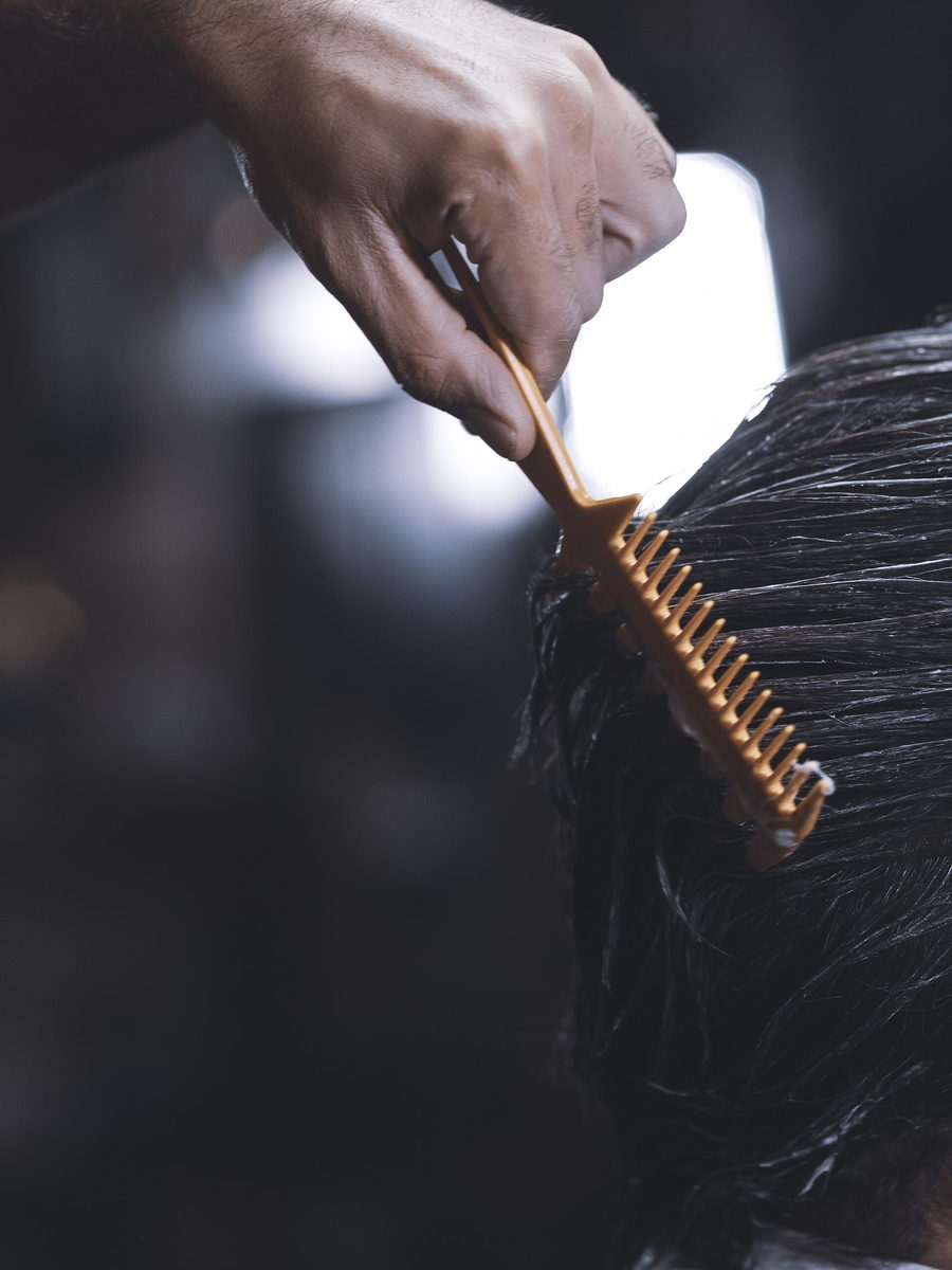 A hand drawing a wide wooden comb through wet dark hair, lit from one side against a deep unlit background.