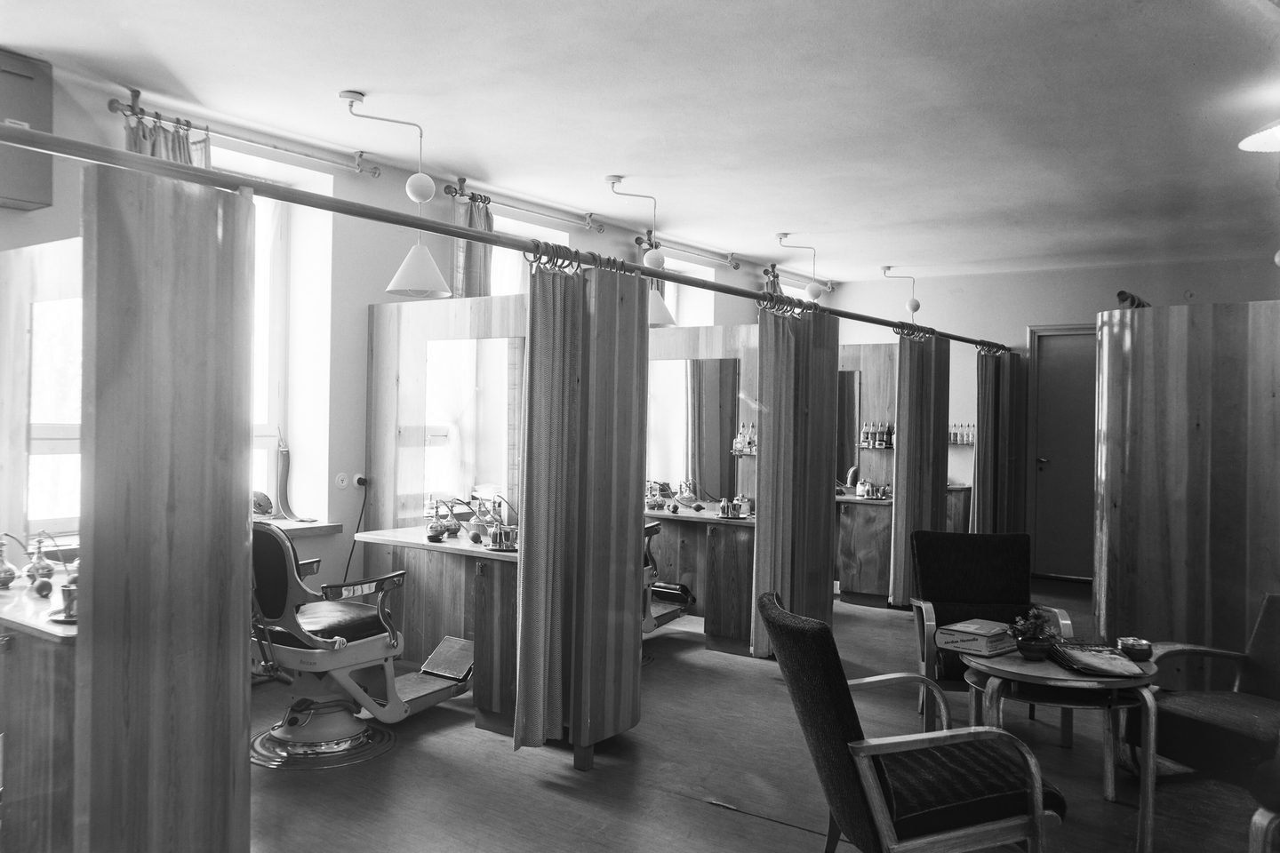A nineteen-forties salon room: a styling chair at a panelled dressing station, with curtained booths along the wall.
