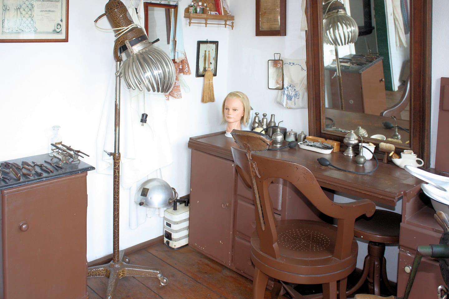 Two chrome hood dryers beside a dressing table holding a blonde practice head.