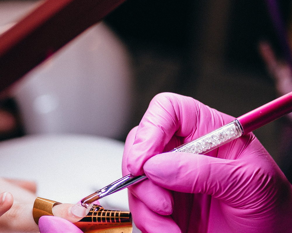 A technician in pink gloves painting a nail with a fine brush, hands only in the frame.