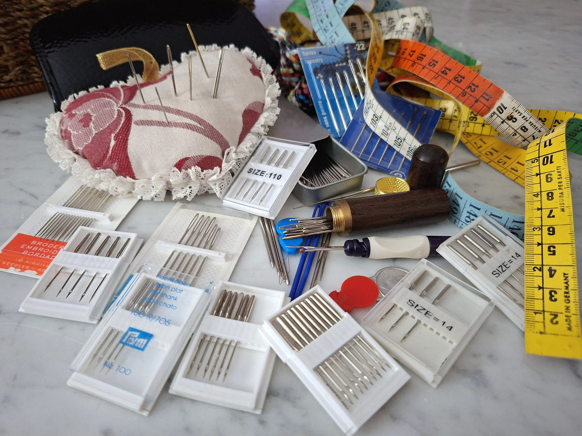 Hand sewing needles, a coiled tape measure and a pincushion laid out on a pale marble workbench, photographed from directly above.