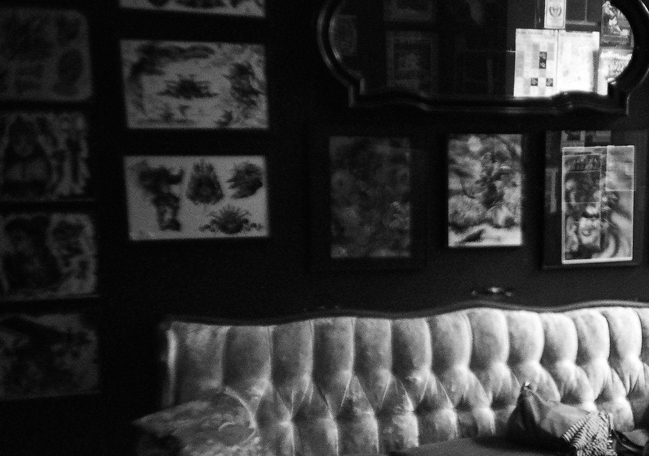 A monochrome tattoo studio interior: a wall of framed flash designs above the buttoned sofa clients wait on.