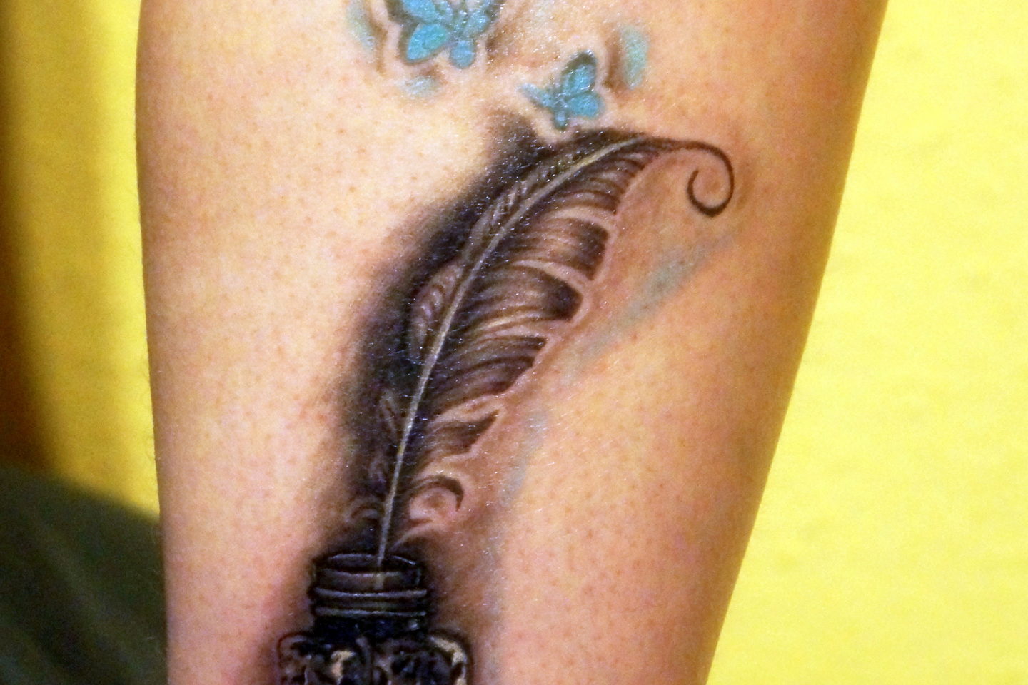 A finished feather-and-inkwell tattoo in black and grey with small blue moths above it.