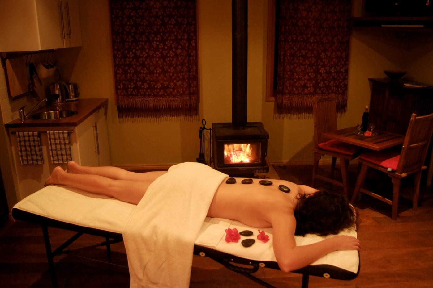 A person lying face down on a massage table with warm stones along the back and red flowers beside them, in a warm room with a lit wood stove.