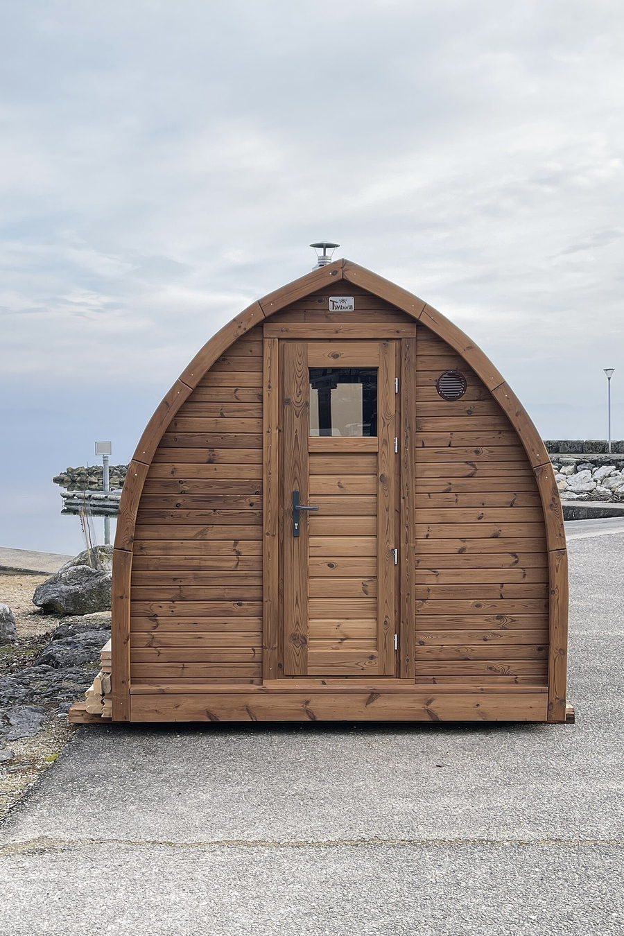 A cedar barrel sauna standing on a stone shore with its door shut and the water behind it.