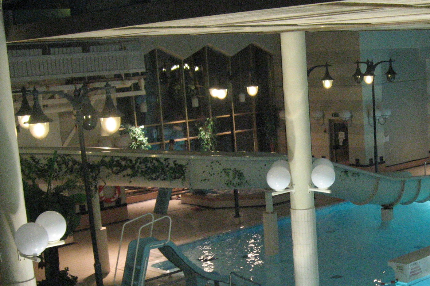 An indoor leisure pool at night, with globe lamps on white columns, a water slide and plants trailing along a balcony.