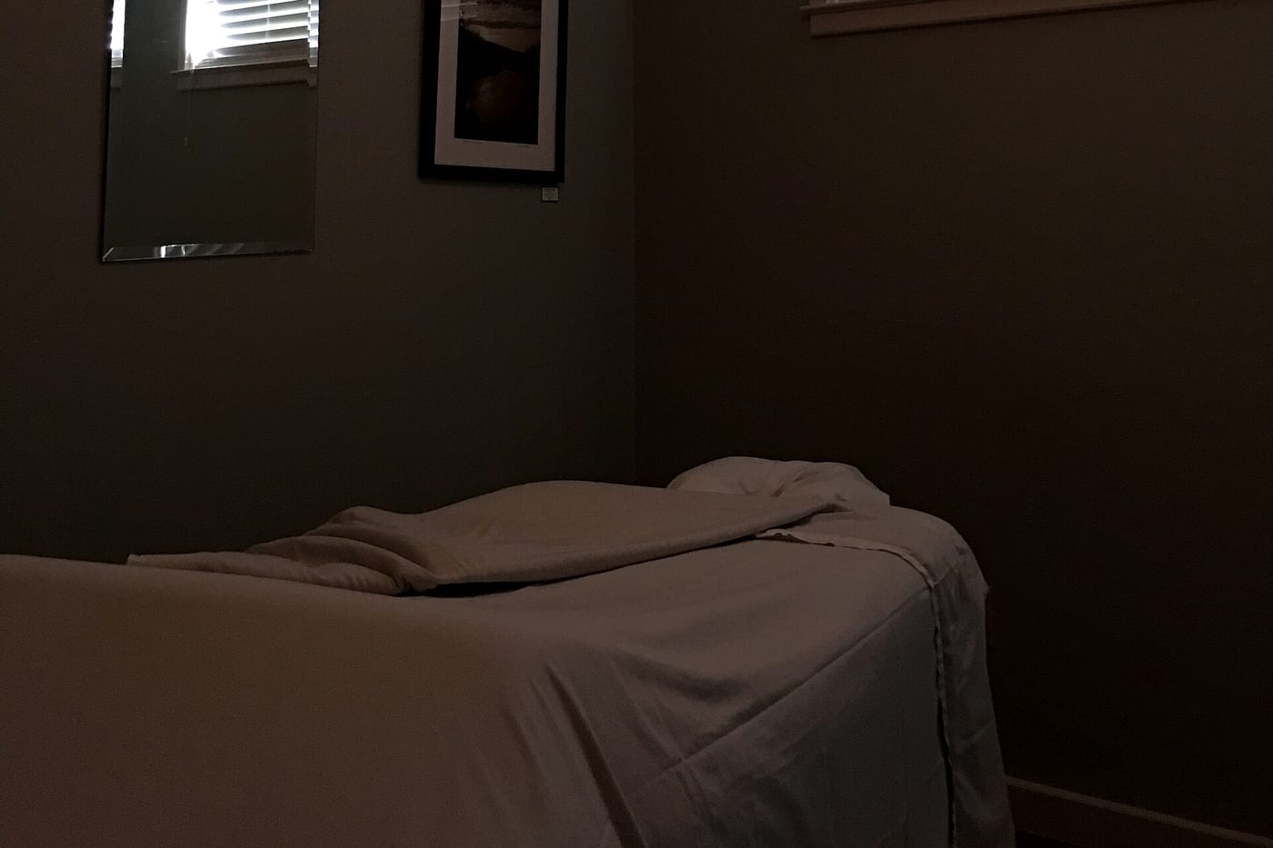 A dim treatment room with a massage table under a grey blanket and a framed picture on the wall.