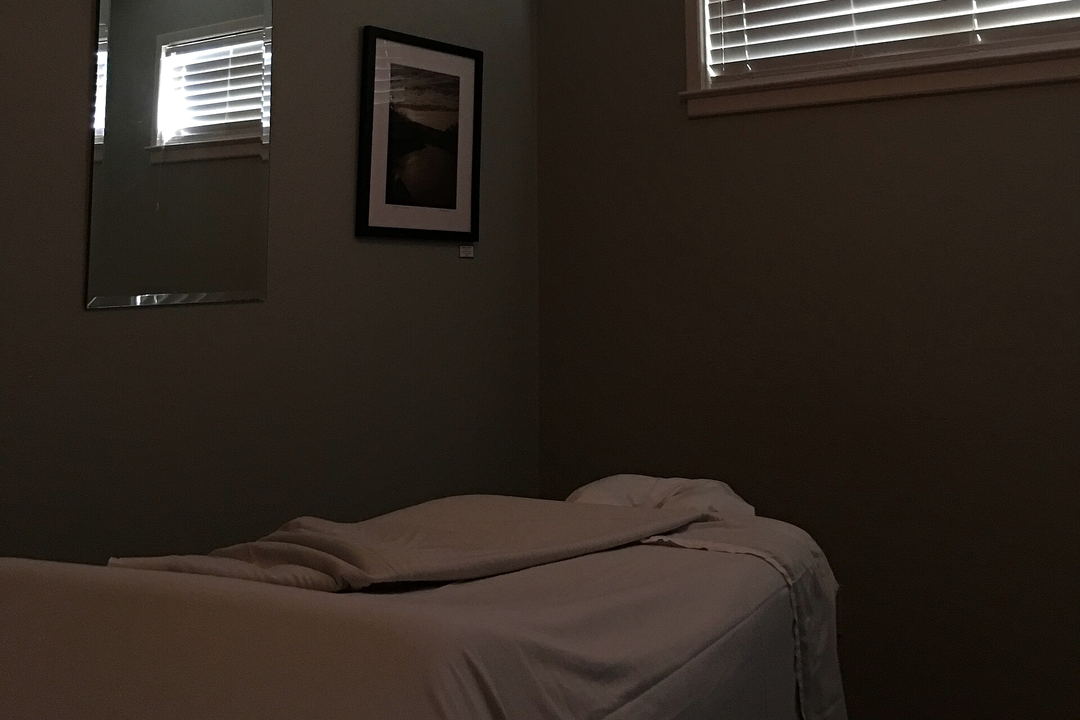 A dim grey treatment room with a made-up massage table, a folded blanket, a framed photograph, a tall mirror and a window with white blinds.