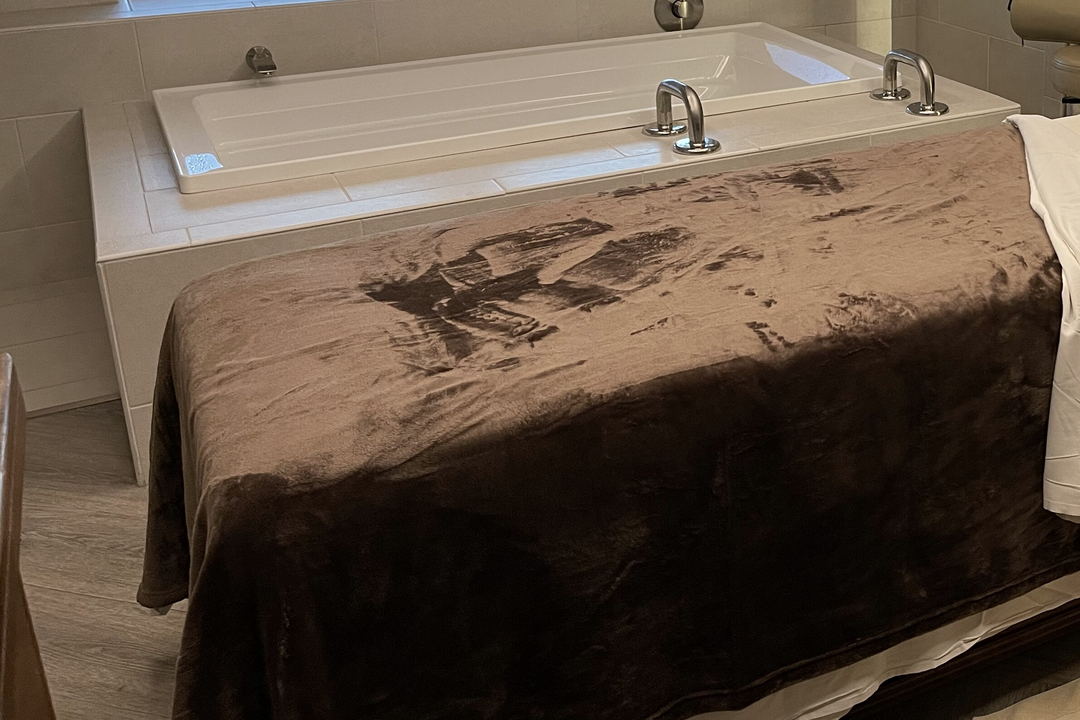 A deep white bathtub with chrome taps beside a treatment table draped in a brown velvet throw.