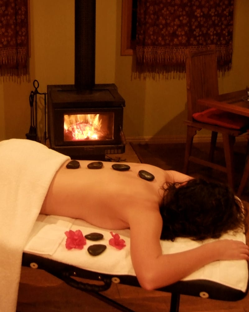 Warm dark stones laid along the back of a person lying face down on a massage table, with a wood stove lit behind.