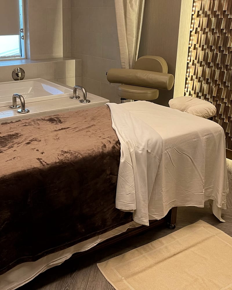 A massage table made up with a white sheet and a brown throw beside a deep bathtub and a wood-tile wall.