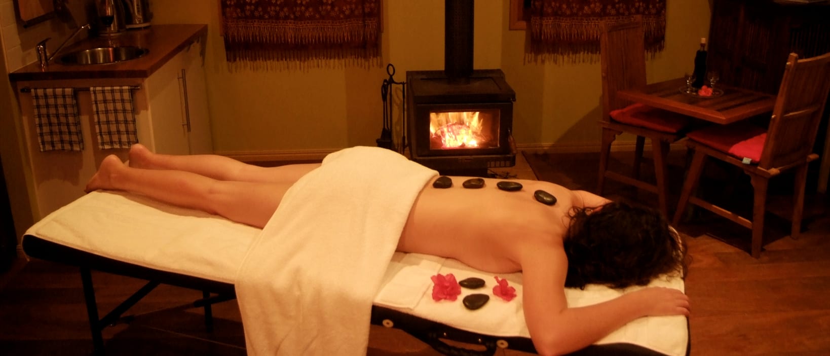 A warm stone massage under way, with dark stones laid along the back of a person lying face down and a wood stove lit behind.