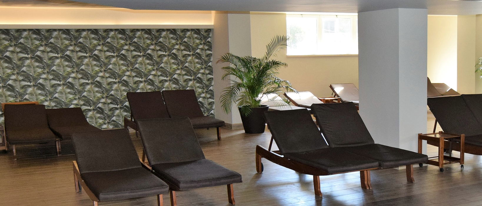 A lounge with rows of dark loungers under palm-leaf wallpaper, with white columns and a slatted wood ceiling.