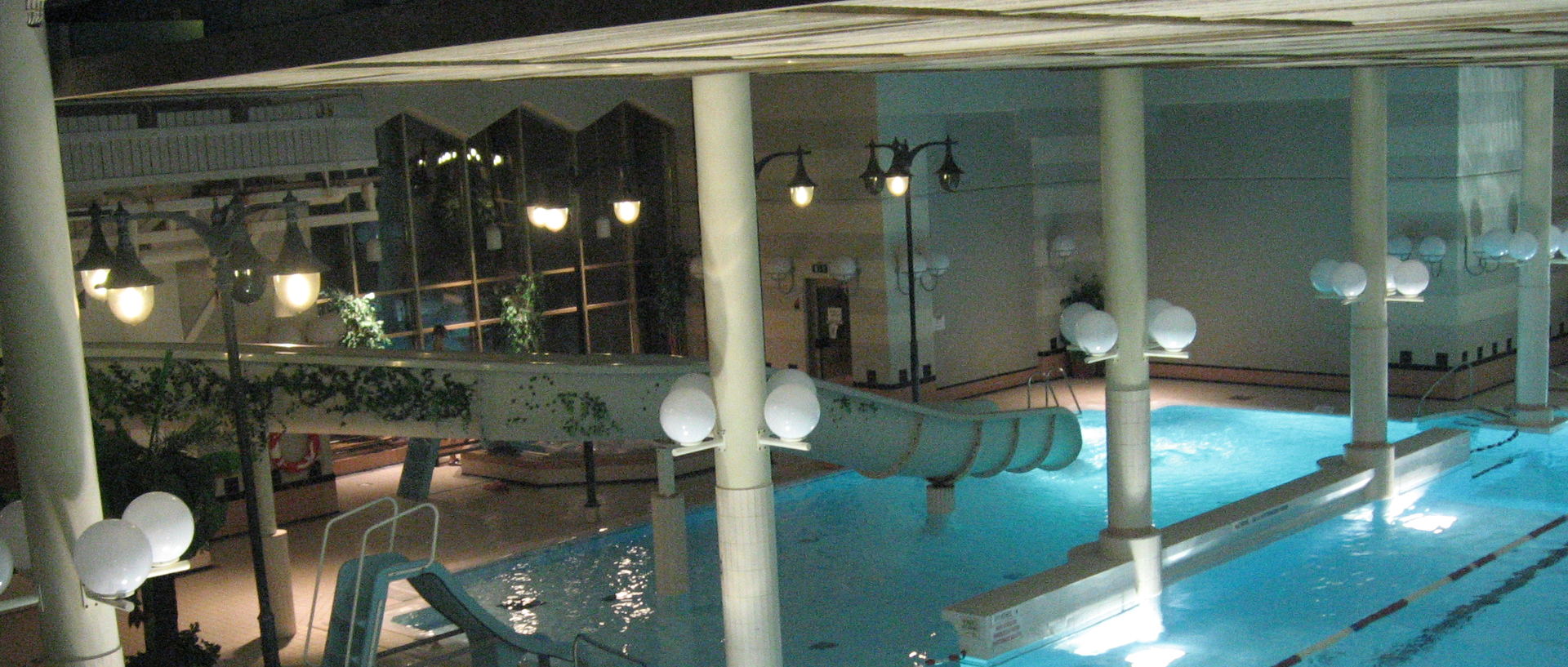 An indoor pool at night under a long canopy, with globe lamps on white columns and a curling water slide.