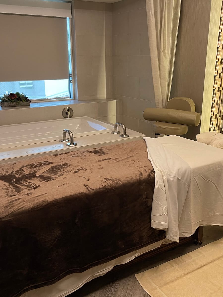 A treatment table made up in white linen beside a corner tub, under a shuttered window.
