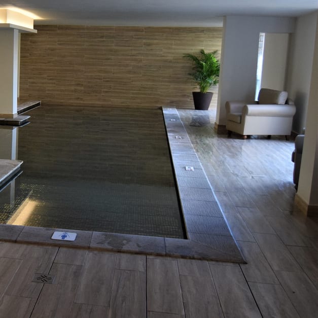 An indoor pool against a wood-tiled wall, with a potted palm and an armchair on the timber floor beyond it.