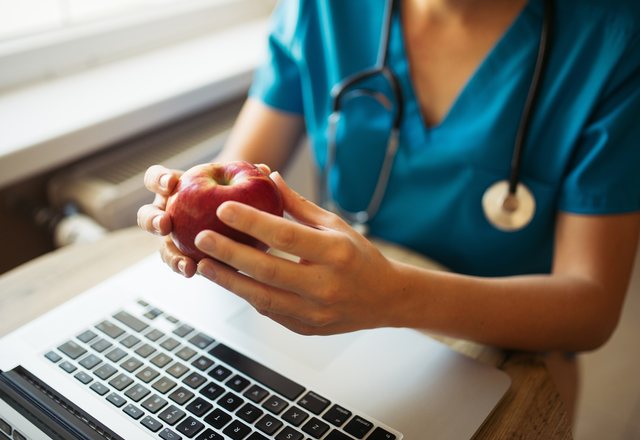 A clinician in scrubs holding an apple at a laptop beside a window