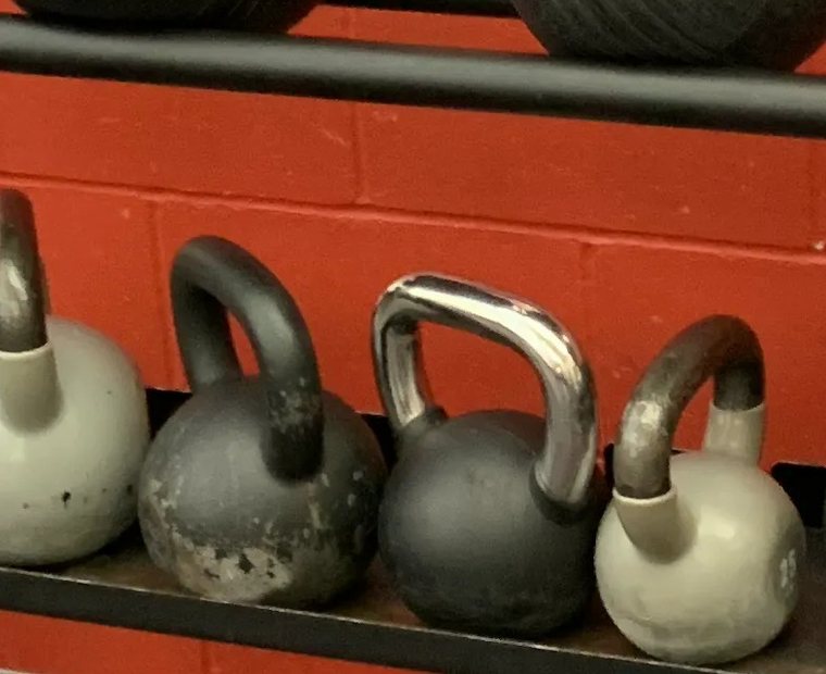 A row of cast-iron kettlebells lined up on the lower shelf of a storage rack against a red block wall