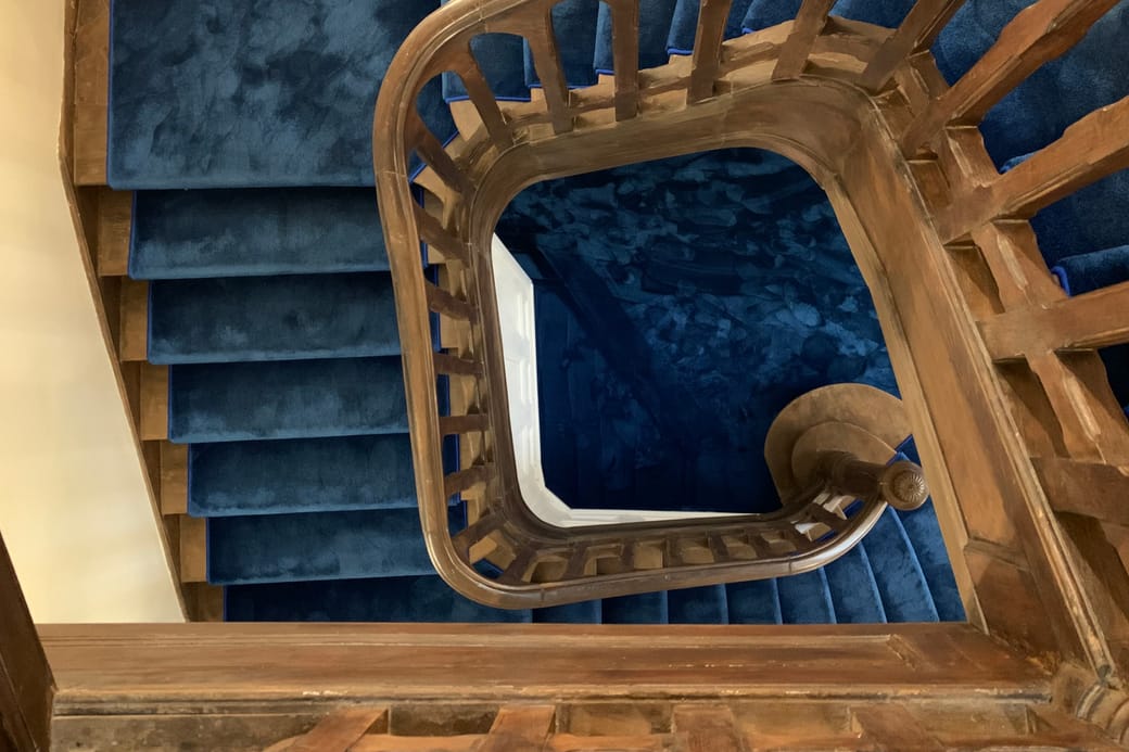 A stairwell seen from above, a wooden balustrade winding around flights of blue carpet.