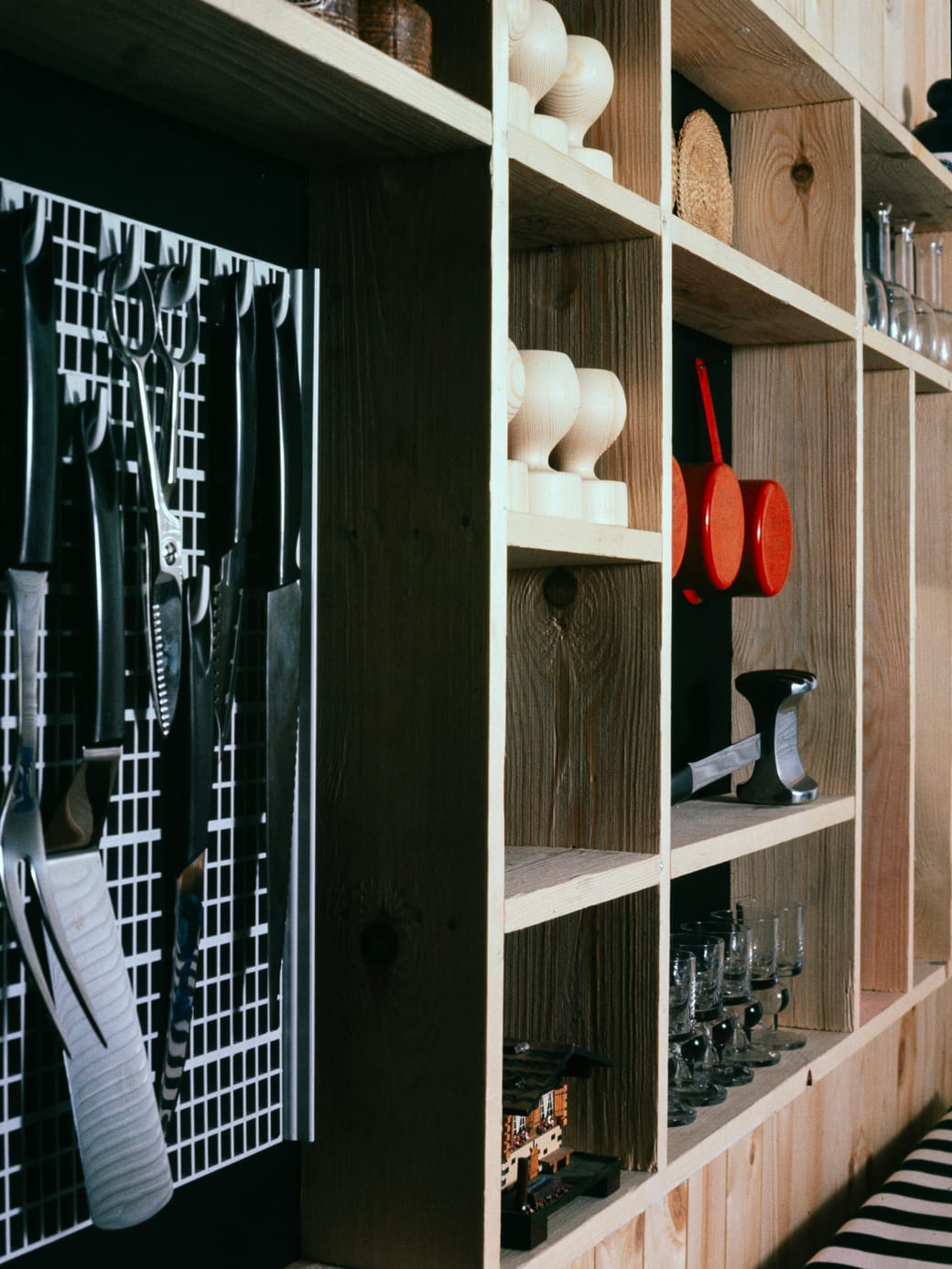 Open pine shelving in a kitchen holding glassware, a red pan and a rack of hanging tools.