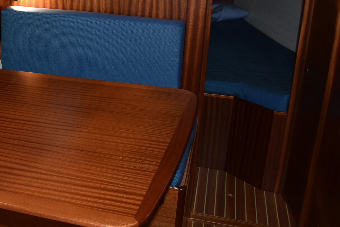 The corner of a yacht saloon, with a mahogany table, blue seat cushions and a berth through the doorway beyond.