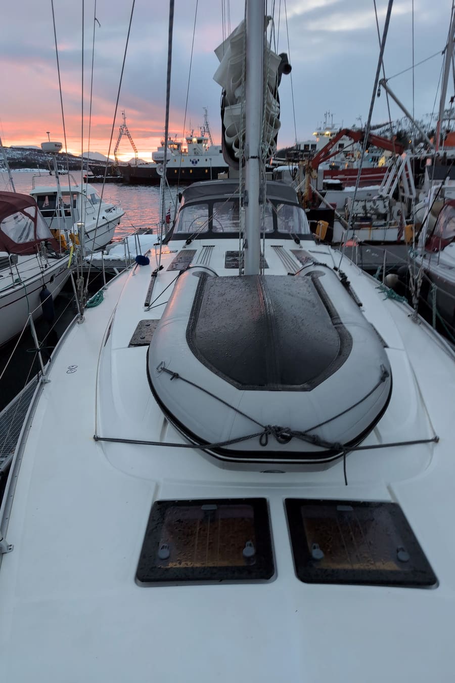 The foredeck of a sailing yacht moored in a marina at dusk, with a deck hatch and the mast rising into the evening sky.