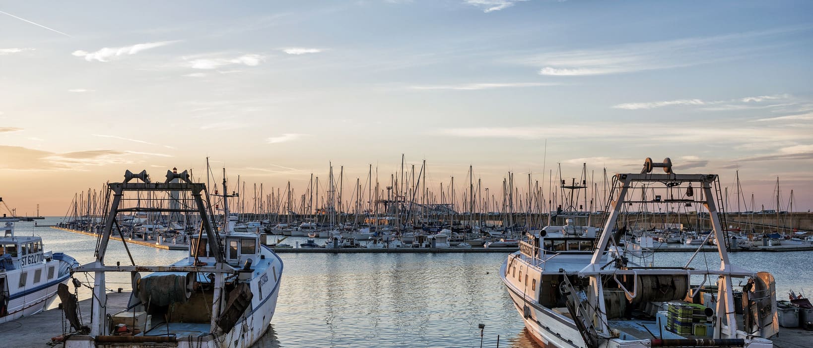Two working boats moored at a marina pontoon at dusk, with rows of yacht masts across the water behind.