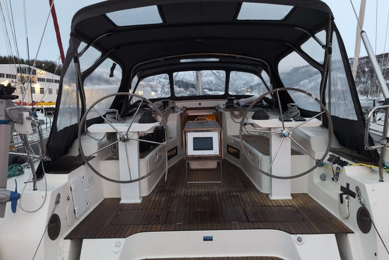 Twin wheels in the cockpit of a sailing yacht under a fitted sprayhood, with a teak sole and a marina beyond.