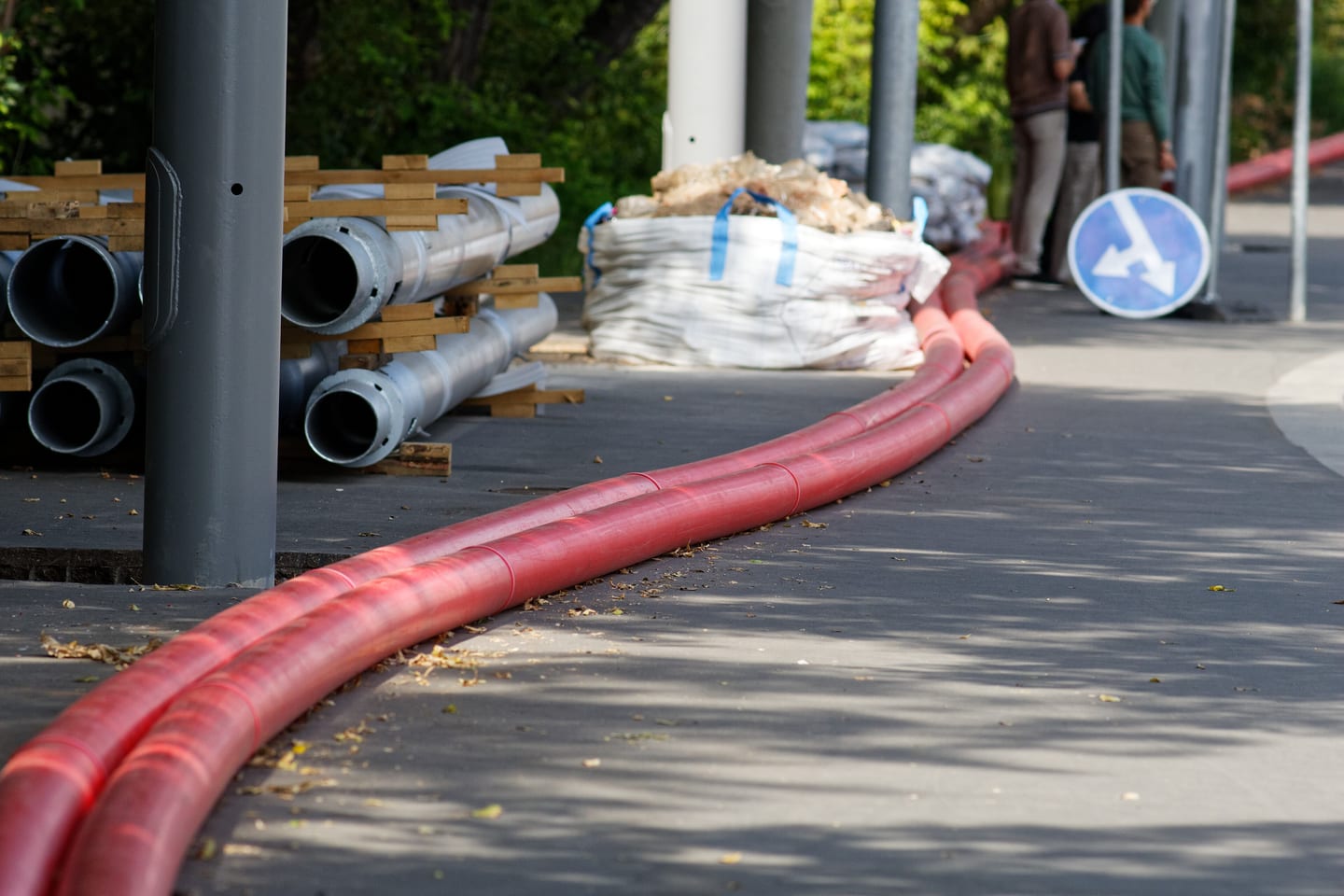 Red cable ducting laid in a curve along a footway beside stacked pipe and bagged material.