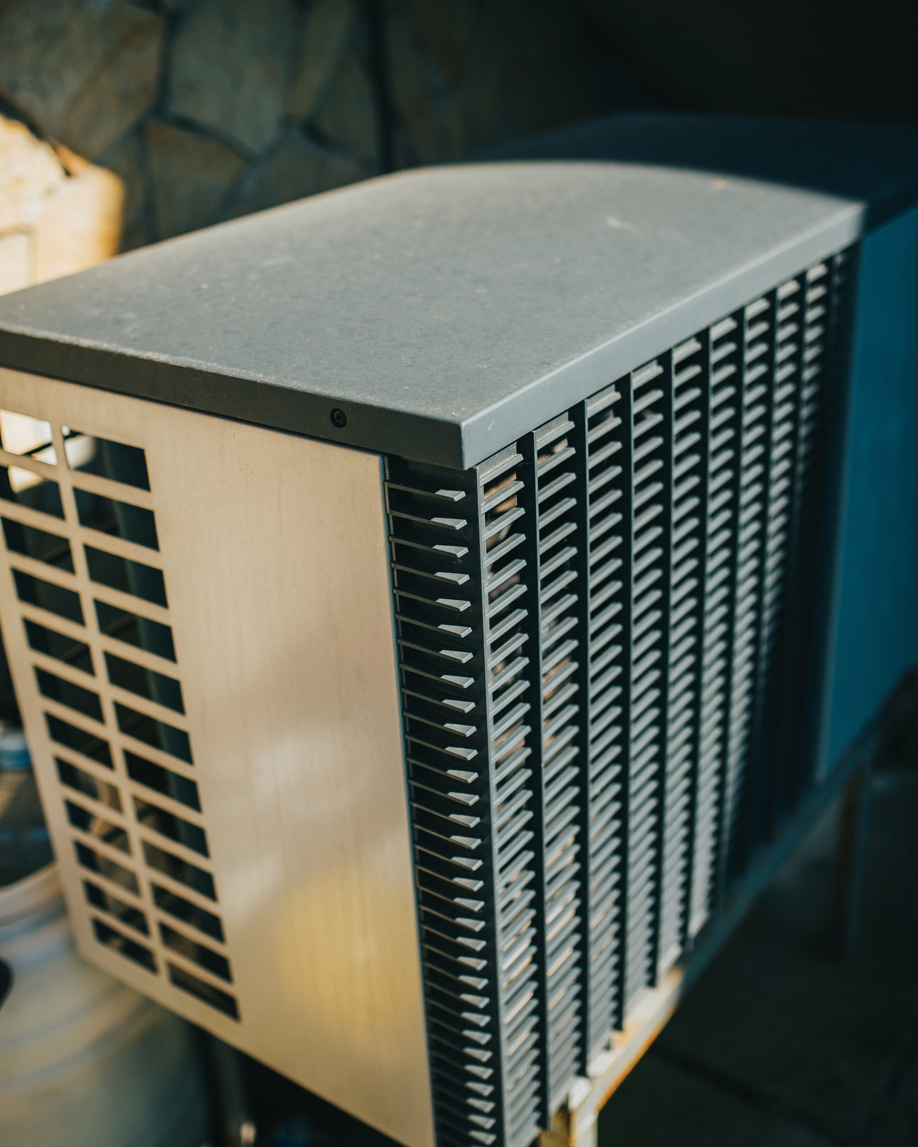 An outdoor condenser unit standing beside a stone wall, its finned coil face turned to the garden.