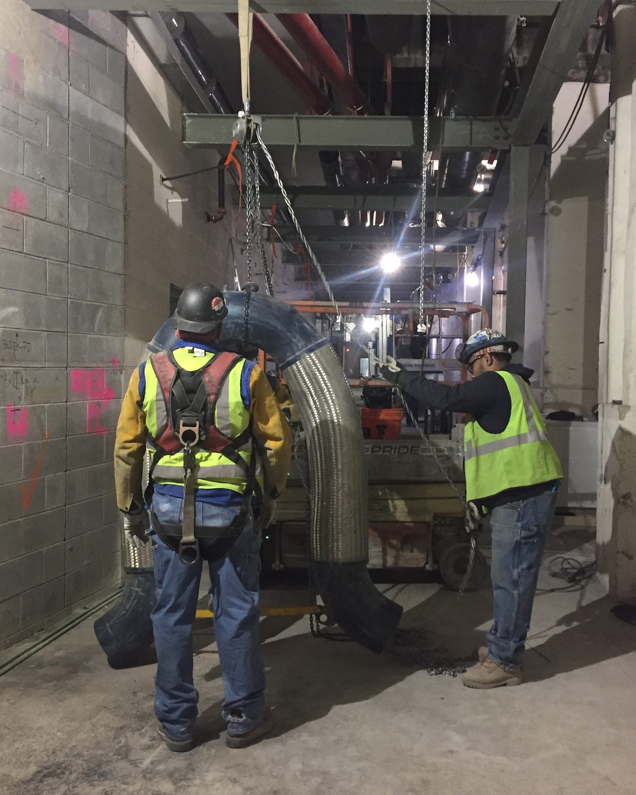 Two technicians in high-visibility vests rigging a large duct elbow into position on a working site.