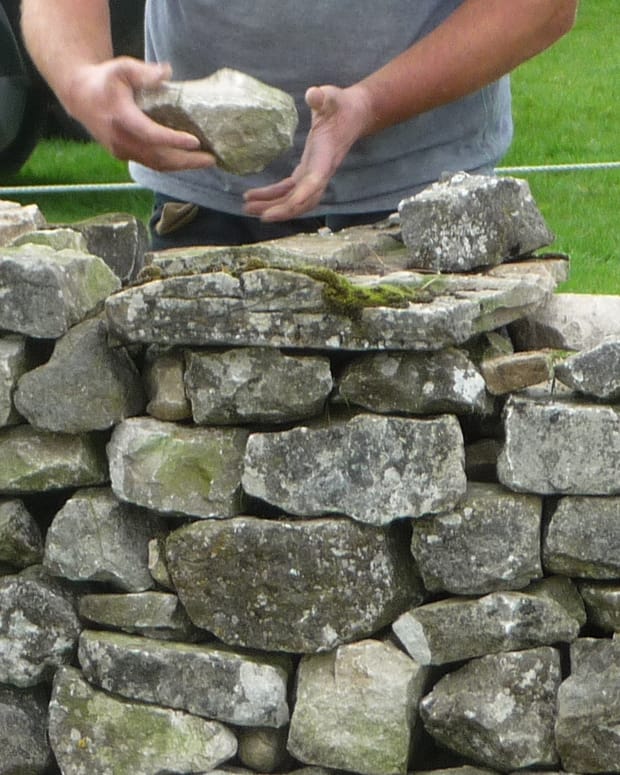 Two hands lifting a piece of fieldstone onto a dry stone wall, cropped at the chest so no face is visible.