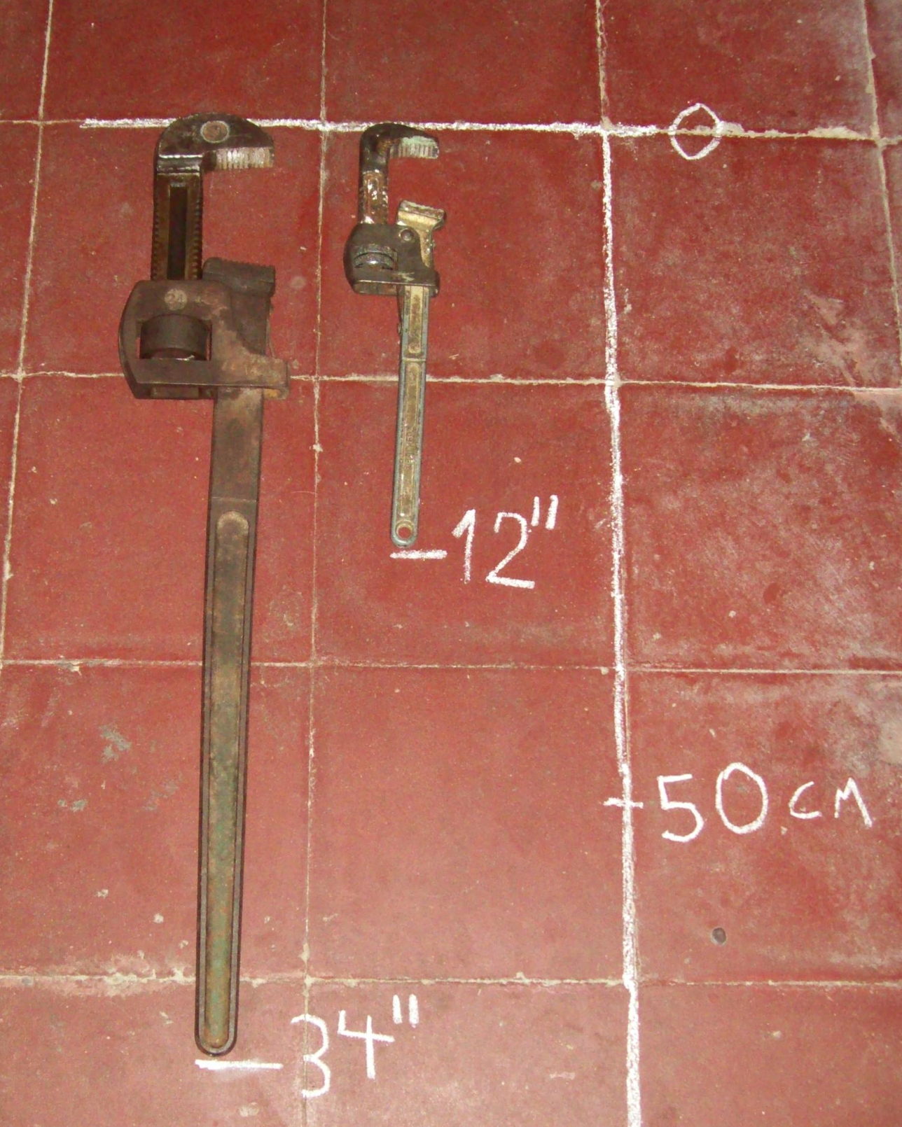 Two pipe wrenches of different sizes laid side by side on a red quarry-tiled floor