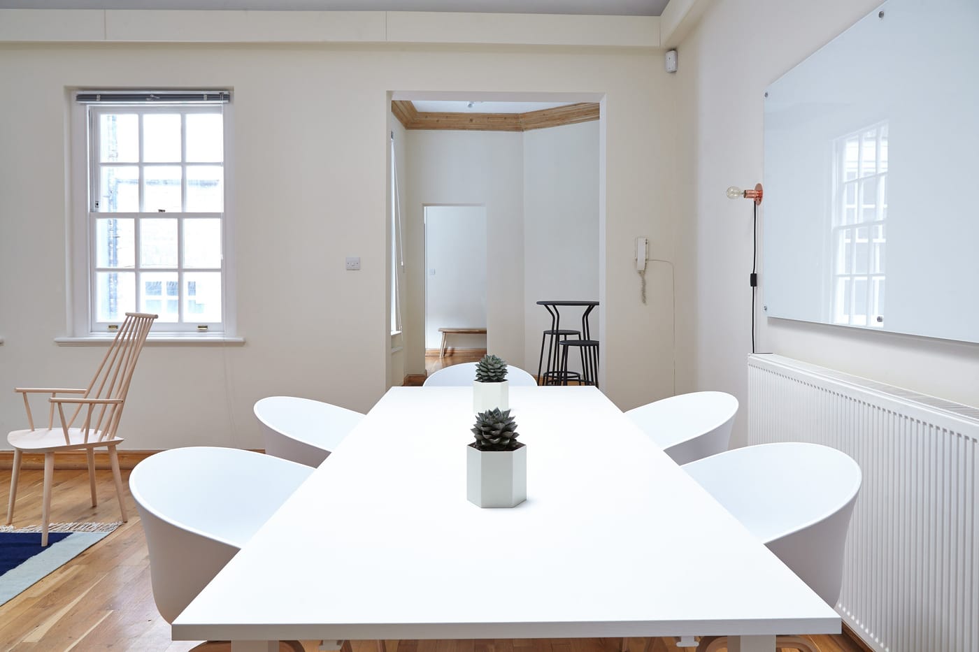 A long white table with white chairs beside a sash window, a glass board and a small succulent.