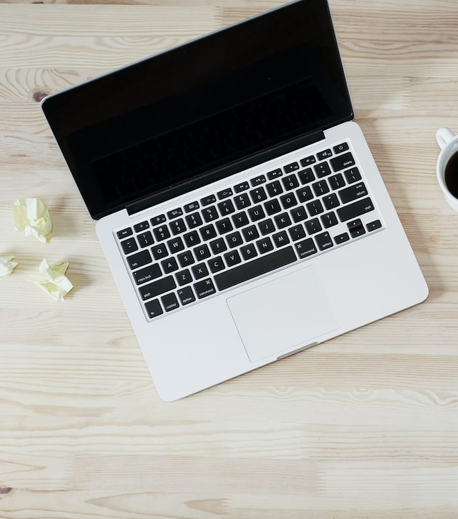 A silver laptop on a pale wooden desk seen from above, with crumpled paper and a coffee cup.