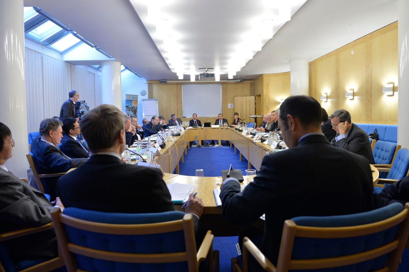 Delegates seen from behind around a U-shaped meeting table with blue chairs.