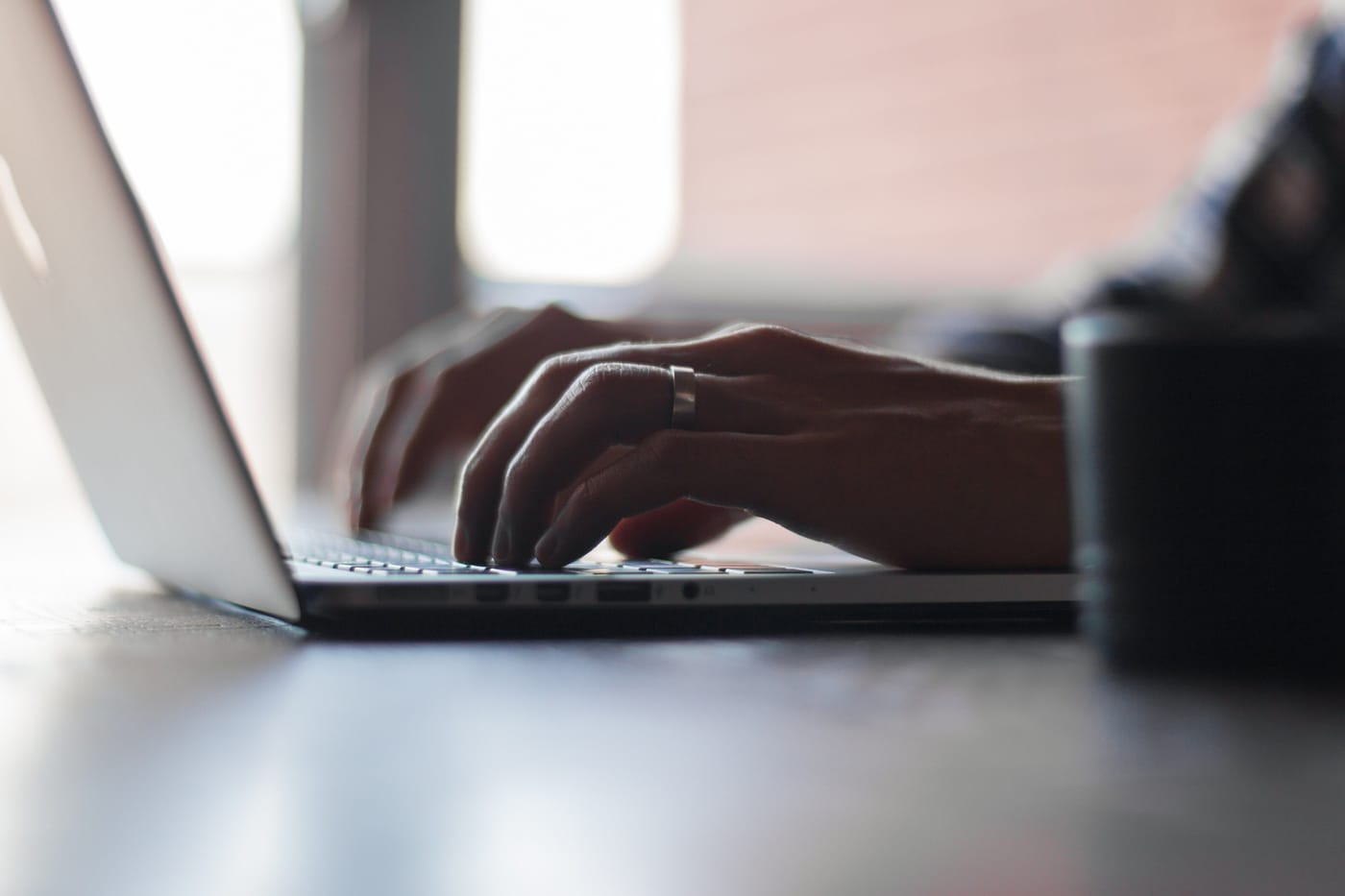 Hands typing on a laptop in window light, beside a dark mug.
