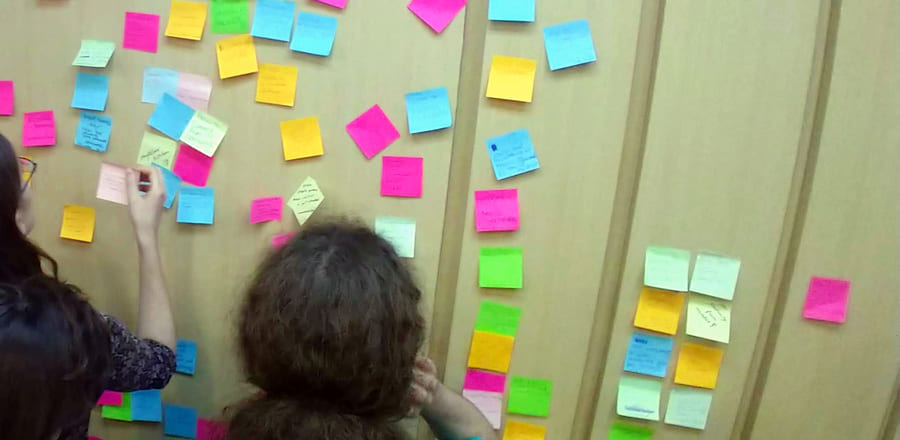 Two people seen from behind adding sticky notes to a wall already covered with them during a workshop
