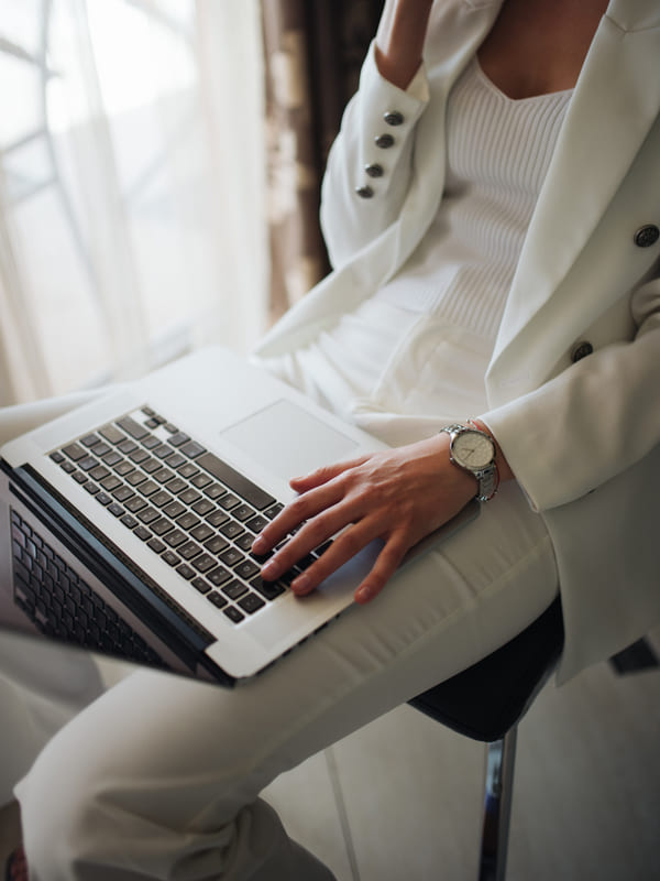 Someone in a light suit working through a laptop resting on their knee beside a window