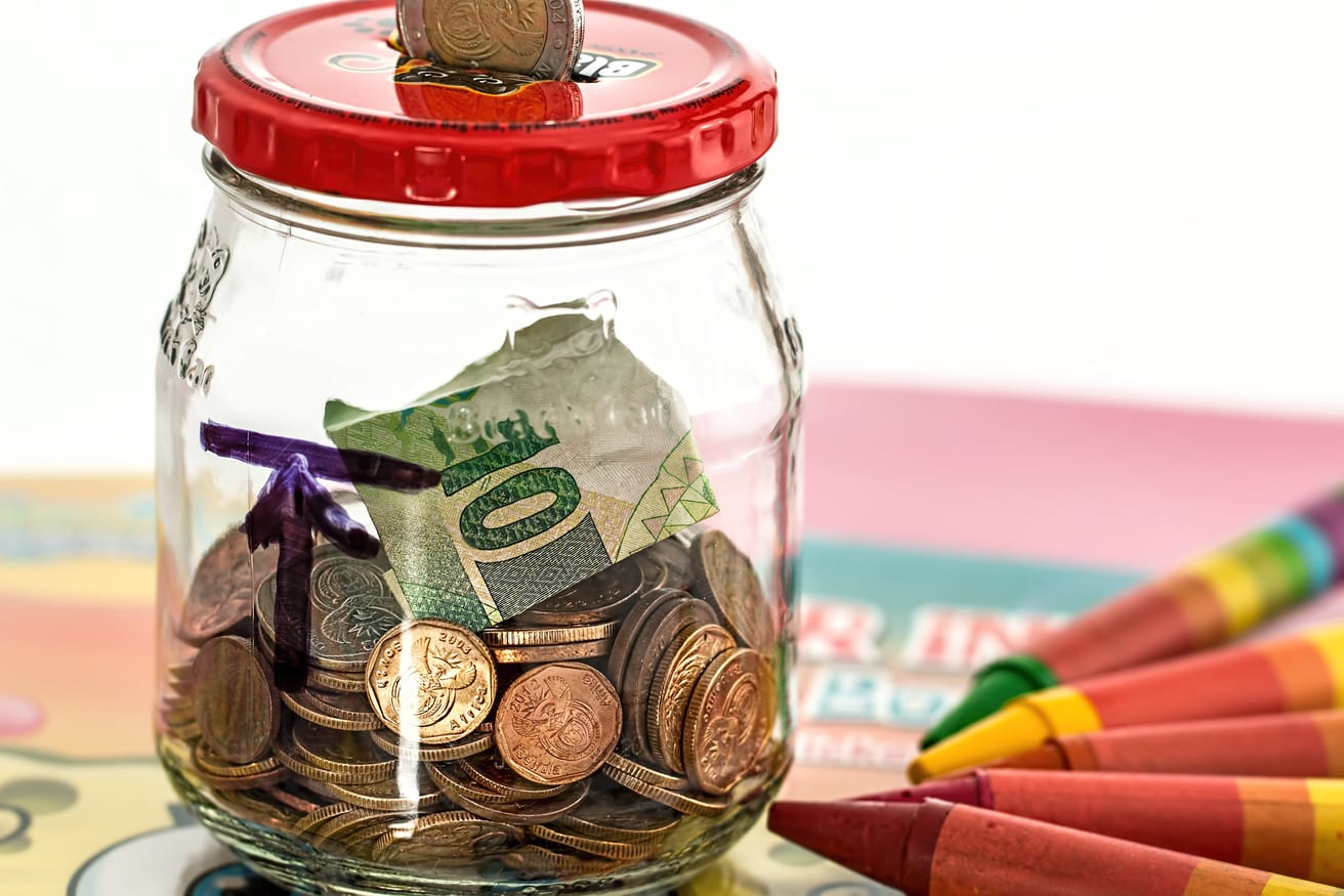 A glass savings jar holding coins, with a handwritten note beside it.