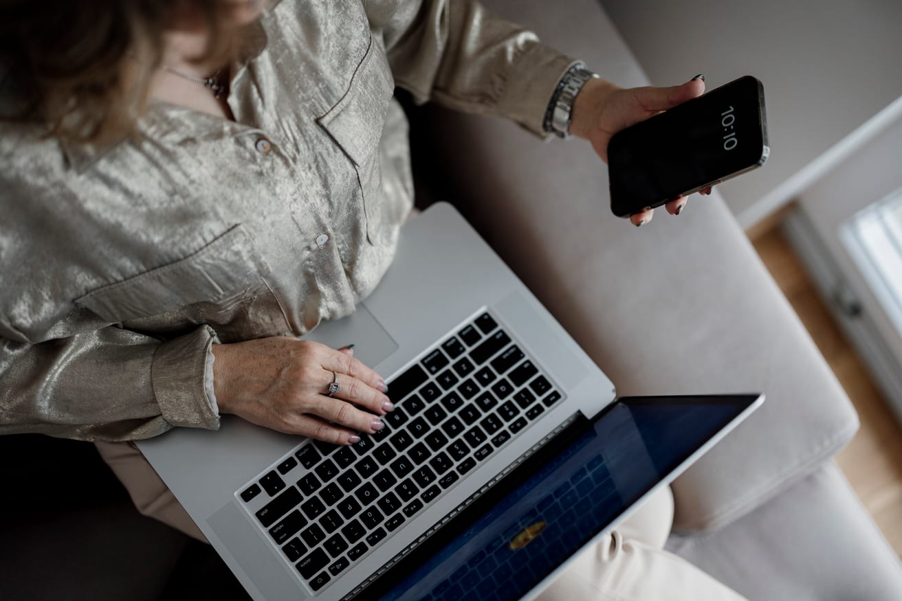 A person working on a laptop while holding a smartphone, face out of frame.