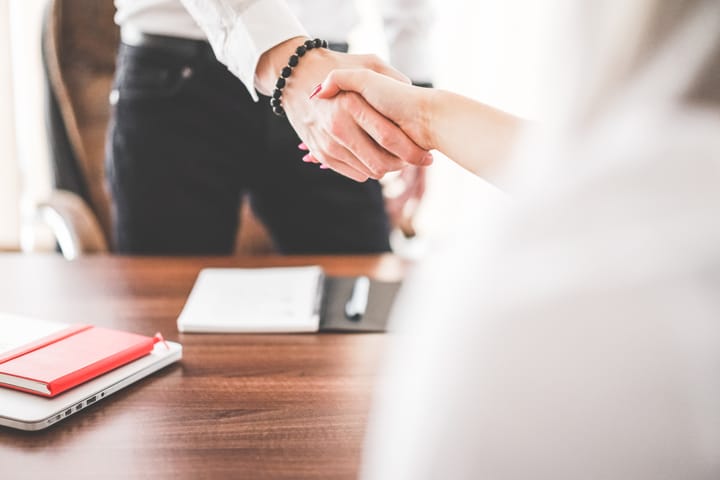Two people shaking hands across a wooden desk holding a notebook and a red folder, their faces out of frame.